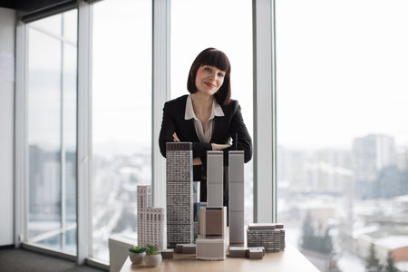 Indoor office portrait of pretty young business lady, wearing white shirt and black suit, standing with arms crossed near 3D megalopolis model in modern office interior with panoramic city view.の写真素材