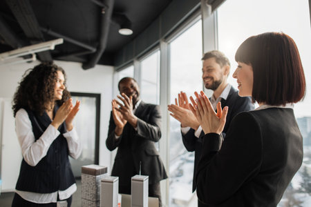 Business negotiations in office room. Team of successful international corporate workers standing near table with model of city district and applauding for successful business strategy of companyの写真素材