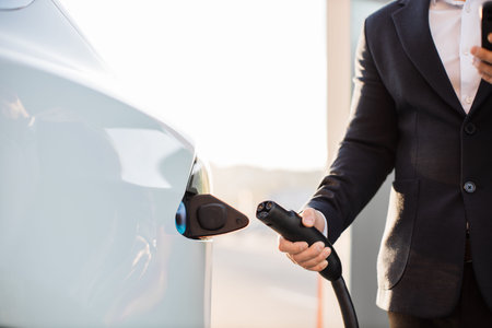 Crop of male hand inserting charging cable into white electric car at outdoors power station. Blurred background. Eco friendly transport with conscious owner.の写真素材