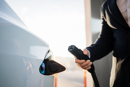 Crop of male hand inserting charging cable into white electric car at outdoors power station. Blurred background. Eco friendly transport with conscious owner.の写真素材