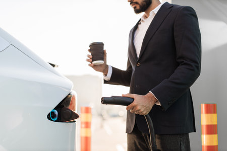 Close up of unrecognizable businessperson in stylish wear standing near white electric car with power cord and coffee cup in hands. EV charging station at urban area.の写真素材