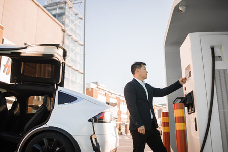 Side view of successful asian businessman paying with credit card for energy at charging station. Stylish man standing outdoors near luxury electro car and using cashless payment.の写真素材