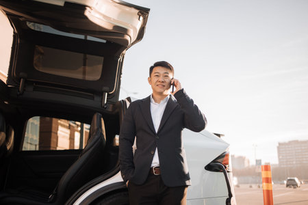 Positive asian man in stylish business wear talking on mobile phone while standing near electric car at outdoors charging station. Blurred background of urban area.の写真素材