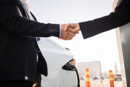 Close up of caucasian woman and asian dealership seller shaking hands in front of white luxury electric car. Concept of people, good deal and new transport.の写真素材