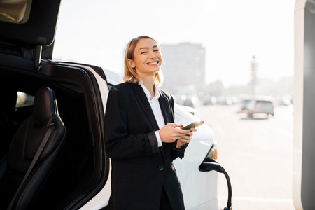 Pretty young lady in business suit cheerfully smiling on camera while using smartphone near modern electric car. Pleasant female blonde standing at EV station for charging her luxury auto.の写真素材