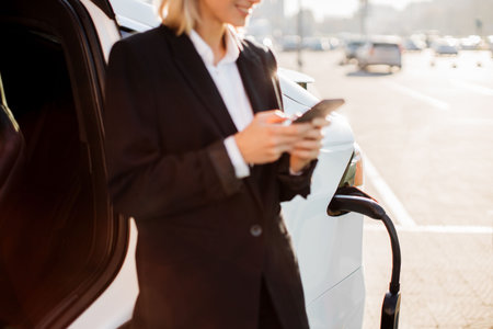 Close up of pleasant caucasian woman in formal clothes with modern smartphone in hands standing near charging electro car outdoors. Blurred background of urban area.の写真素材
