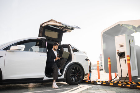 Astylish entrepreneur using wireless laptop while charging electric car on public station. Modern technology for distance work and ecological transport for lifestyles.の写真素材