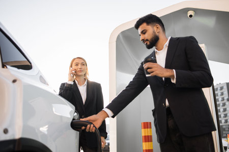 Arabian man in stylish black suit charging white electric car while her caucasian female colleague standing near and talking on mobile phone. Business partners using eco friendly vehicle.の写真素材