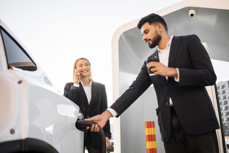 Arabian man in stylish black suit charging white electric car while her caucasian female colleague standing near and talking on mobile phone. Business partners using eco friendly vehicle.の写真素材
