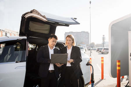 Two focused multiracial business partners in formal attire working on wireless laptop while waiting for luxury electric car to be charged. Concept of people, ecology and technology.の写真素材