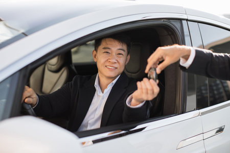 Portrait of pleased asian man in black suit receiving car keys from male dealership seller. Happy male in formal attire buying news luxury vehicle. Concept of successful business and consumerism.の写真素材