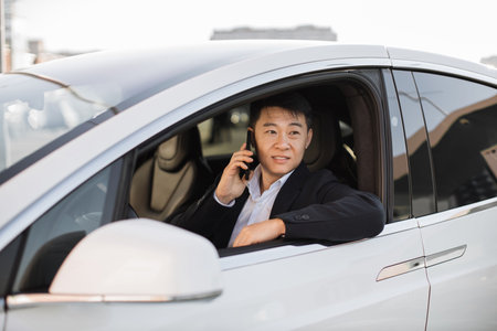 Confident well-dressed businessman having pleasant conversation on mobile phone inside shiny new automobile. Focused asian male thinking over information heard from speaker while staring out window.の写真素材