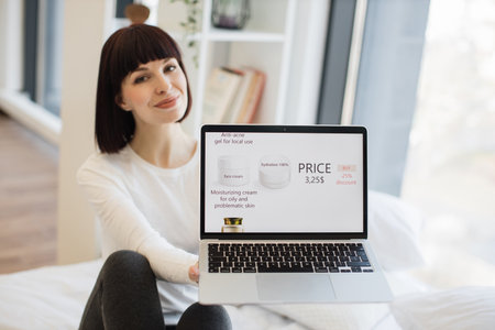 Portrait of beautiful young woman showing modern laptop with cosmetic store on screen. Caucasian brunette in casual wear purchasing face cream and anti-acne gel with 25 percent discount.の写真素材