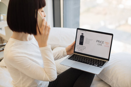 Happy caucasian woman resting on bed with wireless laptop and using smartphone for ordering cosmetic during sale season. Discount of product with 65 percent in online shop.の写真素材