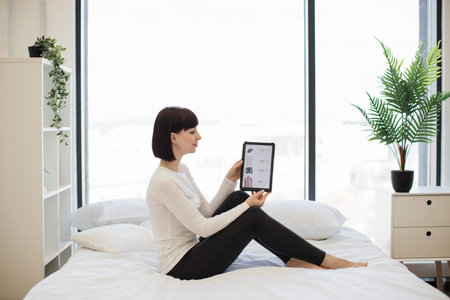 Side view of dark haired caucasian woman scrolling online website with trendy clothes on sale using digital tablet. Casually dressed lady enjoying shopping with big discount while staying at home.の写真素材