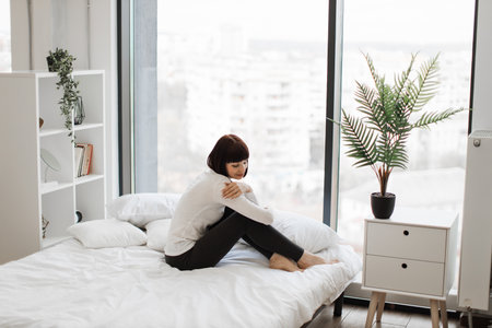 Attractive caucasian woman in domestic outfit resting on white bed with panoramic windows on background. Leisure time at home and lifestyles concept.の写真素材