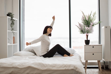 Beautiful woman dressed in domestic clothes stretching arms while sitting on bed with soft white linen. Panoramic windows with city view on background. Dark light at cozy bedroom.の写真素材