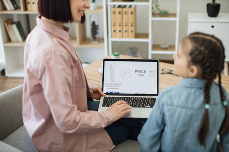 Close up of caucasian woman and little girl sitting on couch with modern laptop and searching available discounts on makeup products. Online store with cosmetics on sale on computer screen.の写真素材