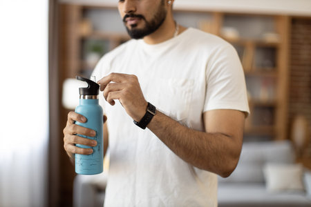 Close up of young arabian man in sport clothes standing at living room and holding bottle of fresh...