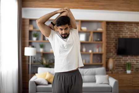 Bearded arabian man in sport outfit warming up with arms stretching at bright living room. Flexible exercises for body care and health condition. Domestic training during free time.の写真素材
