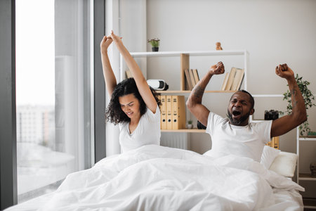 Loving african american couple in casual attire stretching their limbs after night sleep while still lying in bed of bright bedroom. Relaxed family man and woman starting new sunny day in modern flat.の写真素材