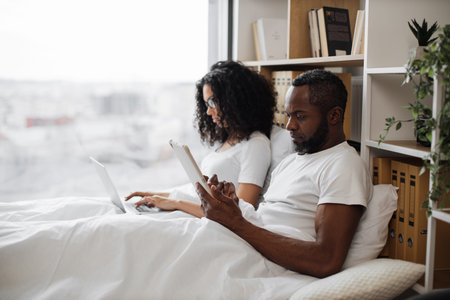 Attractive african american man and pretty woman using digital devices while leaning against soft pillows in bed of studio room. Busy spouses doing their own business without disturbing each other.の写真素材