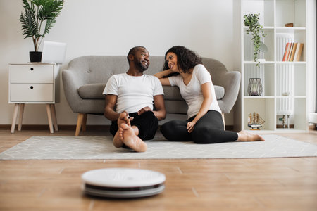 Relaxed multiethnic couple of two lovely people sitting on floor and using remote control for managing work of automated vacuum cleaner. Domestic chores with modern robot.の写真素材