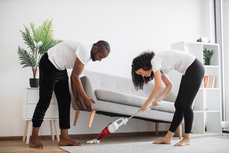 Handsome caring man lifting grey couch for beloved wife for vacuuming underneath. Multicultural young couple in casual clothes cleaning modern apartment together during free time.の写真素材