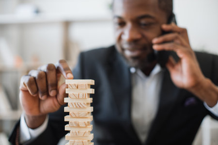 Happy multicultural man in business suit playing while talking on smartphone at office desk with laptop. Cheerful entrepreneur reducing stress at work by enjoying board game during conversation.の写真素材
