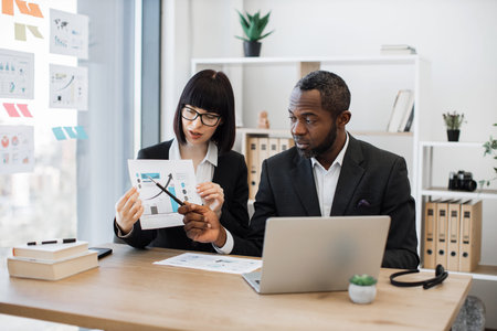 African american man and caucasian woman in business suits being engaged in online briefing while sitting at office desk. Stylish managers presenting information in graphs using portable computer.の写真素材