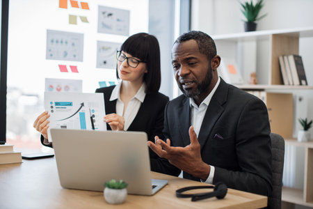 Confident multicultural man communicating online via laptop while attractive caucasian woman demonstrating increase graphs. Efficient business partners discussing project over during video call.の写真素材
