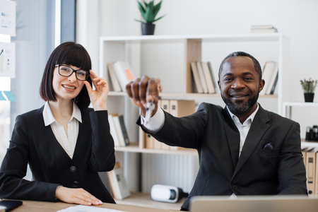 Smiling real estate agents in formal attire holding house keys while sitting at office desk in comfortable workplace. Multiracial man and caucasian woman arranging renting of modern building.の写真素材