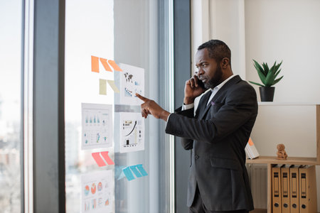 Focused multiracial businessman in smart suit receiving phone call while standing near glass panel with graphs and charts in corner office. Senior executive listening to workers speech over mobile.の写真素材