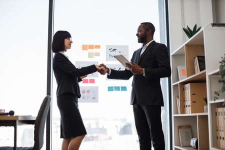 Confident bespectacled woman and smiling man with digital device shaking hands in front of panoramic window of conference hall. Multiracial entrepreneurs sealing agreed business deal at work.の写真素材