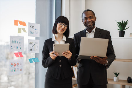 Multiethnic male with portable computer and caucasian female with digital tablet standing in modern workplace. Business colleagues in smart clothes planning new commercial strategy in office.の写真素材