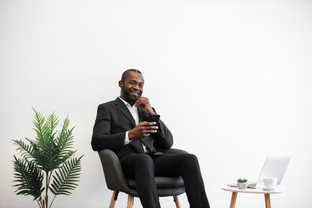 African man in business suit smiling cheerfully on camera while using modern smartphone at office center. Laptop and coffee cup on table near. Concept of people, business and technology.の写真素材