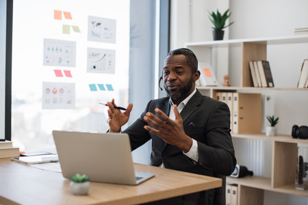 African american man in formal suit having video call at office using headset and wireless laptop. Transparent board with various graphs and charts on background. Concept of online business meeting.の写真素材