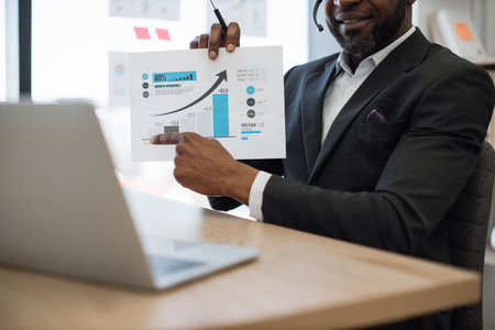 Close up of african american man in headset explaining paper report with graphs during video conference on modern laptop. Office worker in black suit presenting market growth to employees online.の写真素材