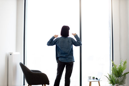 Back view of relaxed female brunette stretching arms while standing at spacious office near panoramic window. Caucasian young employee in casual attire enjoying coffee break during working hours.の写真素材