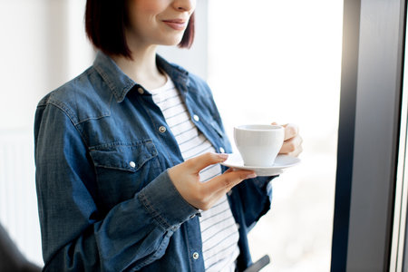 Close up of pleasant caucasian woman in casual wear holding cup of hot beverage while standing at office. Panoramic window on blurred background. Short coffee break before work.の写真素材