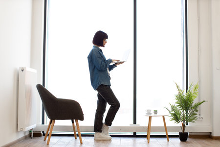 Charming female employee typing on wireless laptop while standing near panoramic window at office. Dark haired woman in casual outfit and glasses checking emails on modern gadget at workplace.の写真素材