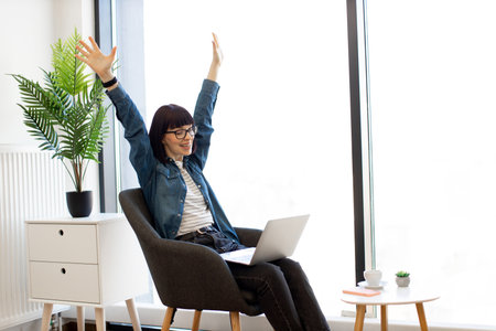 Happy woman in casual clothes showing victory gesture with raised hands while sitting in office chair with laptop on knees. Joyful female expressing excitement from successful deal during video call.の写真素材