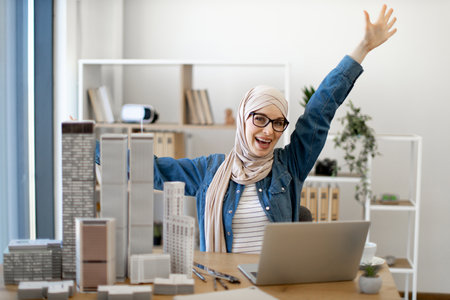 Happy arabian female wearing hijab and spectacles raising arms in victory joy while enjoying work in architecture office. Successful engineer celebrating commercial agreement with winners gesture.の写真素材