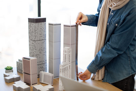 Close up view of architectural models presenting innovative metropolitan area set on office desk indoors. Female architect in casual clothes using metal ruler for measuring multi-storey building.の写真素材