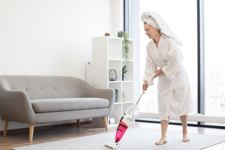 Young woman in bathrobe and towel on head removing dust from soft carpet using cordless vacuum cleaner in spacious living room. Cheerful housewife enjoying housekeeping with modern appliances.の写真素材