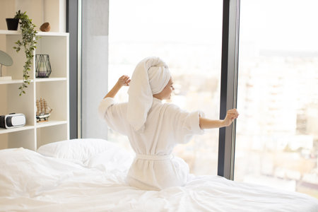 Back view of young lady in bath towel and white robe pulling hands aside while sitting in front of panoramic window in airy bedroom. Cheerful woman relieving tension before starting new day at home.の写真素材