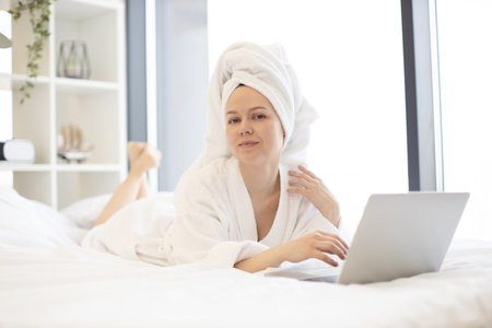 Beautiful young lady in white after-shower robe daydreaming on cozy bed while running portable computer in sunny room of modern apartment. Charming female adult enjoying comfortable lifestyle.の写真素材
