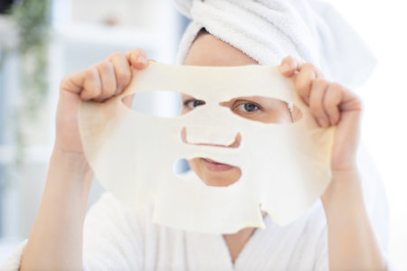 Charming caucasian female in white robe and bath towel looking through holes of cosmetic sheet mask cut out for eyes on background of home interior. Pretty woman having fun with face care product.の写真素材