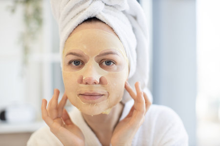 Close up of beautiful calm lady in after-shower wear using cosmetic tissue mask loaded with skincare benefits during at-home spa day. Healthy mature person following weekly facial treatment routine.の写真素材