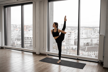 Caucasian dark-haired woman practicing advanced yoga standing in trivikramasana pose in white spacious studio. Professional yogi wearing comfortable sports clothing balancing on one leg doing asana.の写真素材